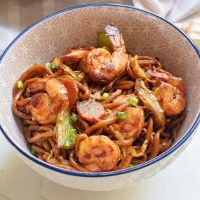 A bowl of stir-fried noodles with shrimp, bell peppers, and sliced green onions, all in a rich, glossy sauce. The noodles and ingredients are mixed together, topped with large pieces of shrimp, and served in a decorative bowl.