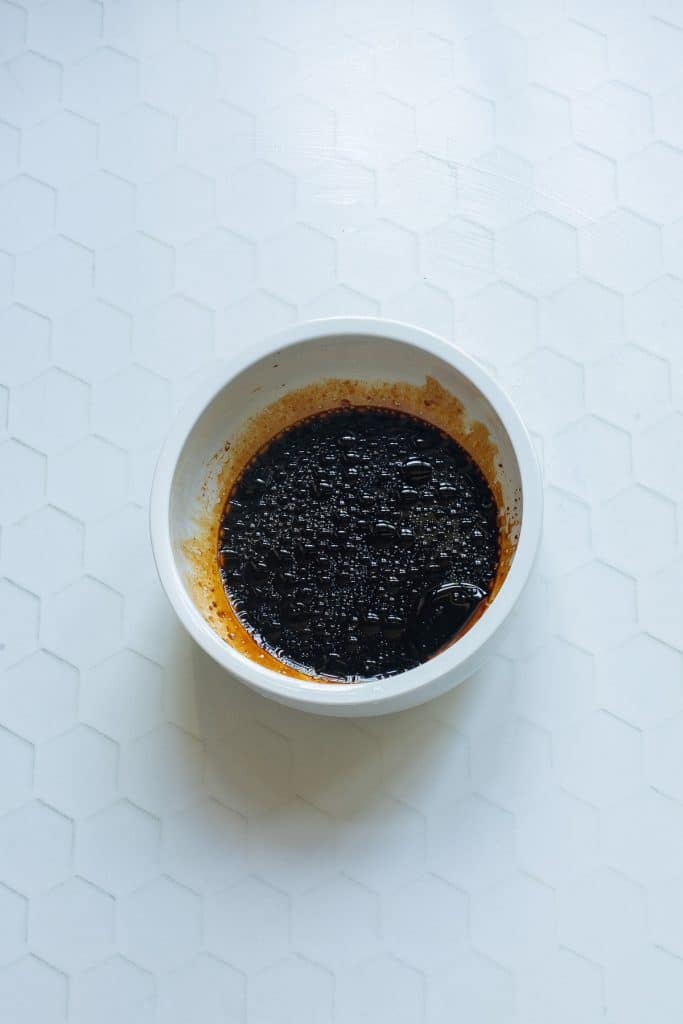 A white bowl filled with a dark, viscous liquid placed on a hexagonal patterned white surface. Small specks are visible in the liquid.