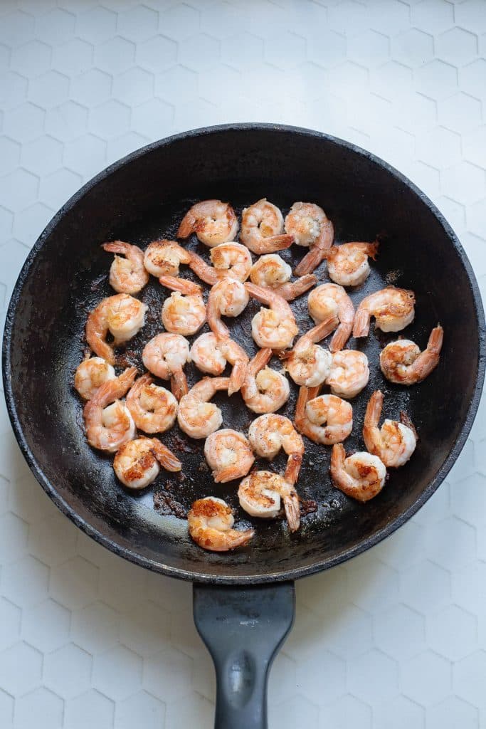 A black frying pan containing cooked shrimp, lightly browned and seasoned, is placed on a white hexagonal tiled surface. The shrimp are evenly distributed across the pan, ready to be served.