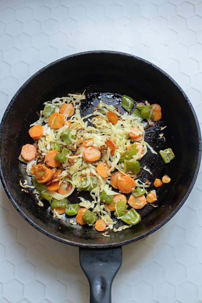 A black skillet on a white hexagonal-patterned surface contains sautéed vegetables, including sliced carrots, bell peppers, and shredded cabbage. The vegetables are lightly cooked and scattered evenly across the pan.