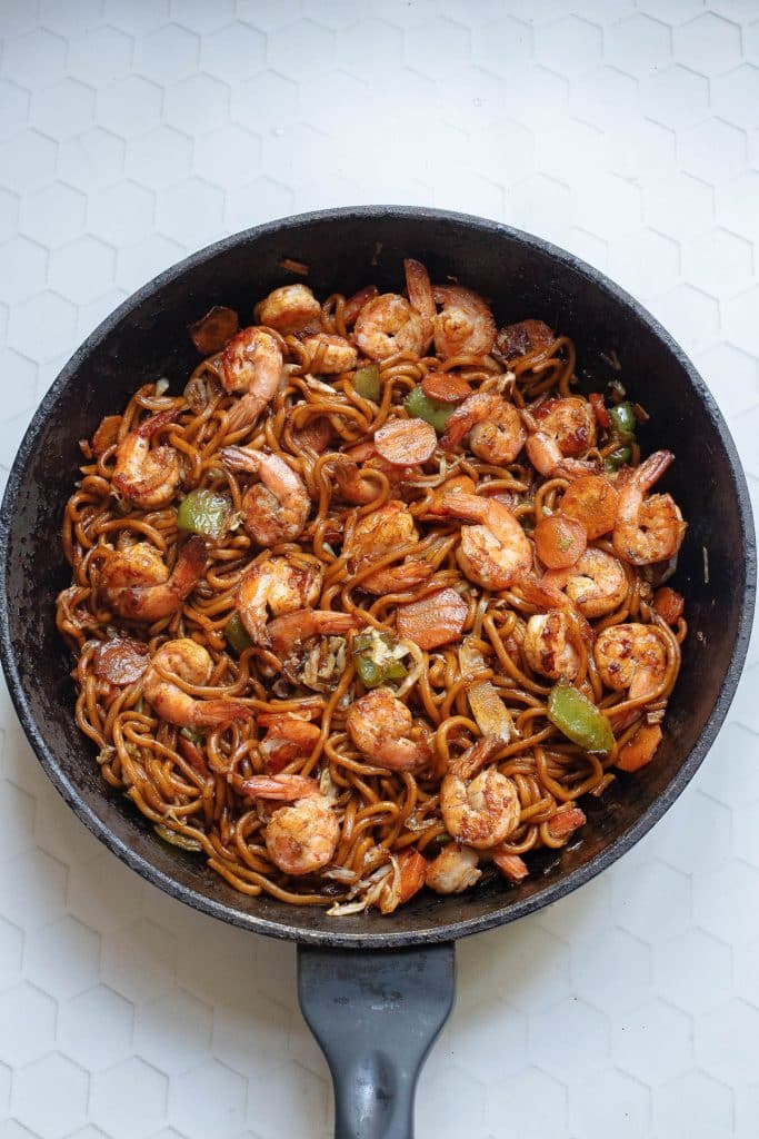 A skillet filled with cooked noodles, shrimp, sliced vegetables, and spices. The mix of ingredients is evenly coated with a rich, savory sauce, creating a vibrant and appetizing dish. The skillet rests on a white, hexagonal-tiled surface.