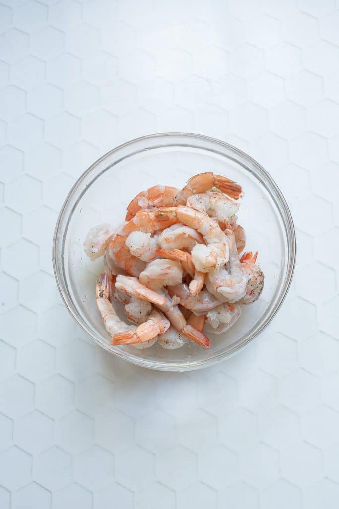 A glass bowl filled with cooked shrimp is placed on a white, patterned surface. The shrimp are peeled and appear ready to be used in a recipe.