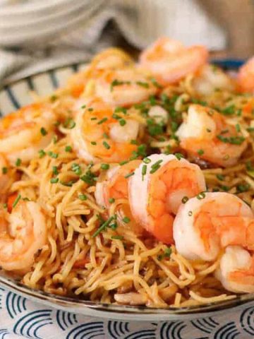A plate of shrimp stir-fried noodles garnished with chopped green onions. The dish is served on a patterned plate, placed on a blue cloth, with a rustic wooden table in the background.