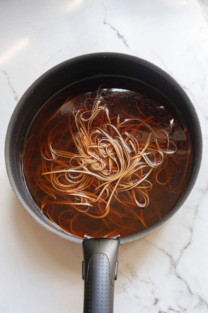 A black saucepan on a marble surface contains thin, long noodles soaking in a dark liquid.