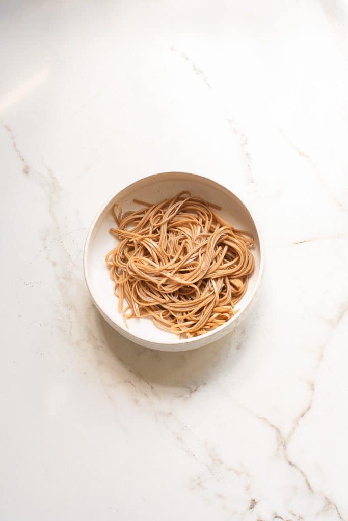 A white bowl filled with cooked spaghetti sits on a white marble surface. The light illuminates the pasta, showcasing its texture.