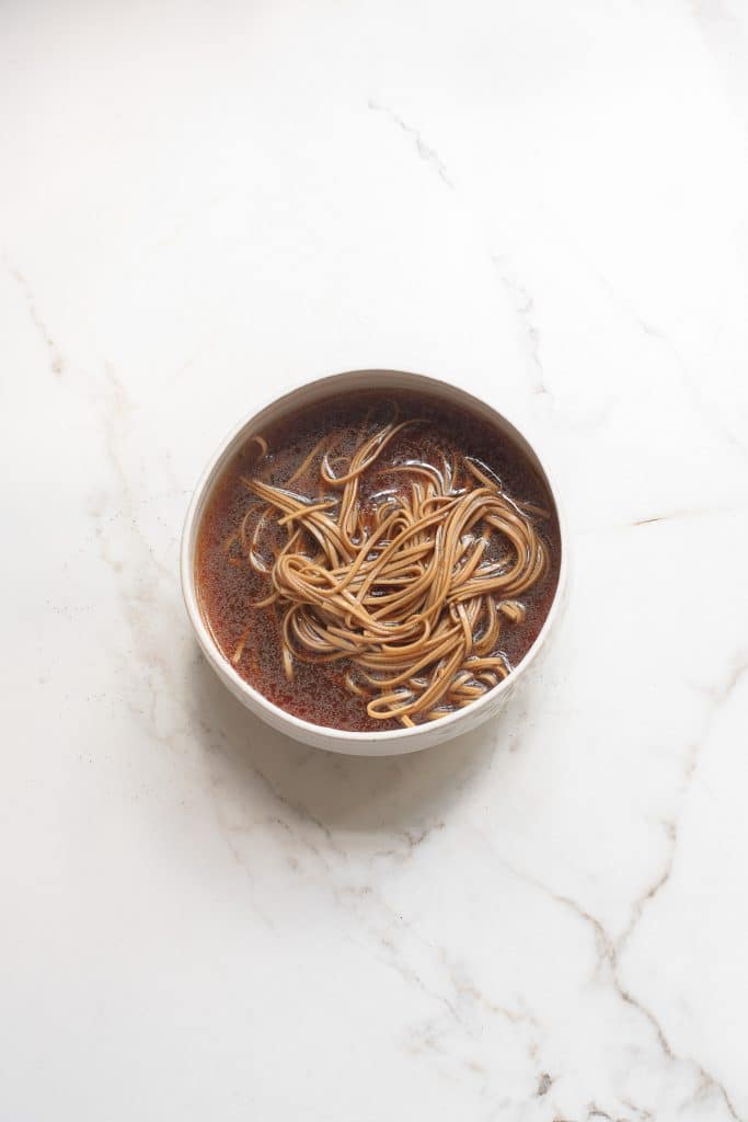A bowl of brown soup with long, thin noodles on a white marble surface. The soup appears seasoned and the noodles are partially submerged, creating a spiral pattern.