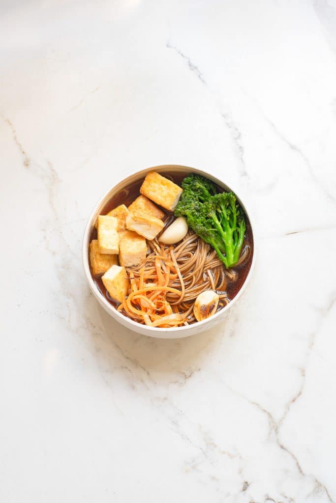 A bowl of soba noodles topped with cubed tofu, broccoli, grated carrot, and garlic on a white marble surface.