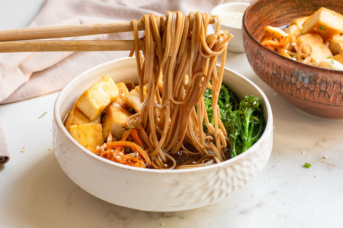 A bowl of noodle soup with chopsticks lifting noodles. The bowl contains tofu cubes, broccoli, and carrots. There's a second bowl in the background on a marble surface.