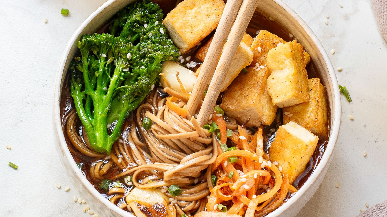 A bowl of soba noodles in broth, topped with broccolini, crispy tofu, sautéed mushrooms, spiralized carrots, and sprinkled with sesame seeds. Wooden chopsticks rest on top of the tofu.