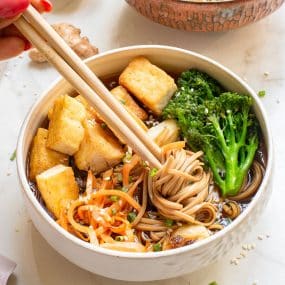 A bowl of soba noodles with chopsticks, featuring tofu cubes, broccolini, and shredded carrots. The dish is garnished with sesame seeds and chopped green onions.