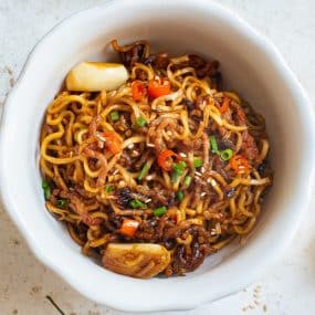 A white bowl filled with stir-fried noodles, garnished with chopped green onions, red chili slices, sesame seeds, and pieces of garlic on a light-colored surface.