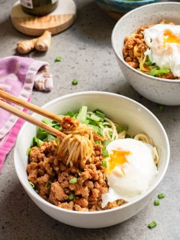 A bowl of noodles topped with minced meat, chopped green onions, and a poached egg with a runny yolk. Chopsticks rest on the bowl. In the background, another bowl and ingredients like ginger and leafy greens are visible on a gray countertop.