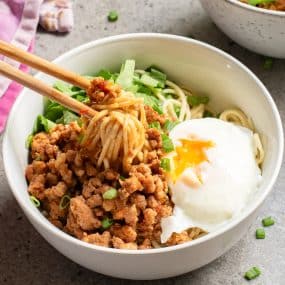 A white bowl filled with noodles, topped with minced meat, a poached egg with a runny yolk, and sliced greens. A pair of chopsticks is lifting some noodles from the bowl. The bowl is placed on a gray countertop.