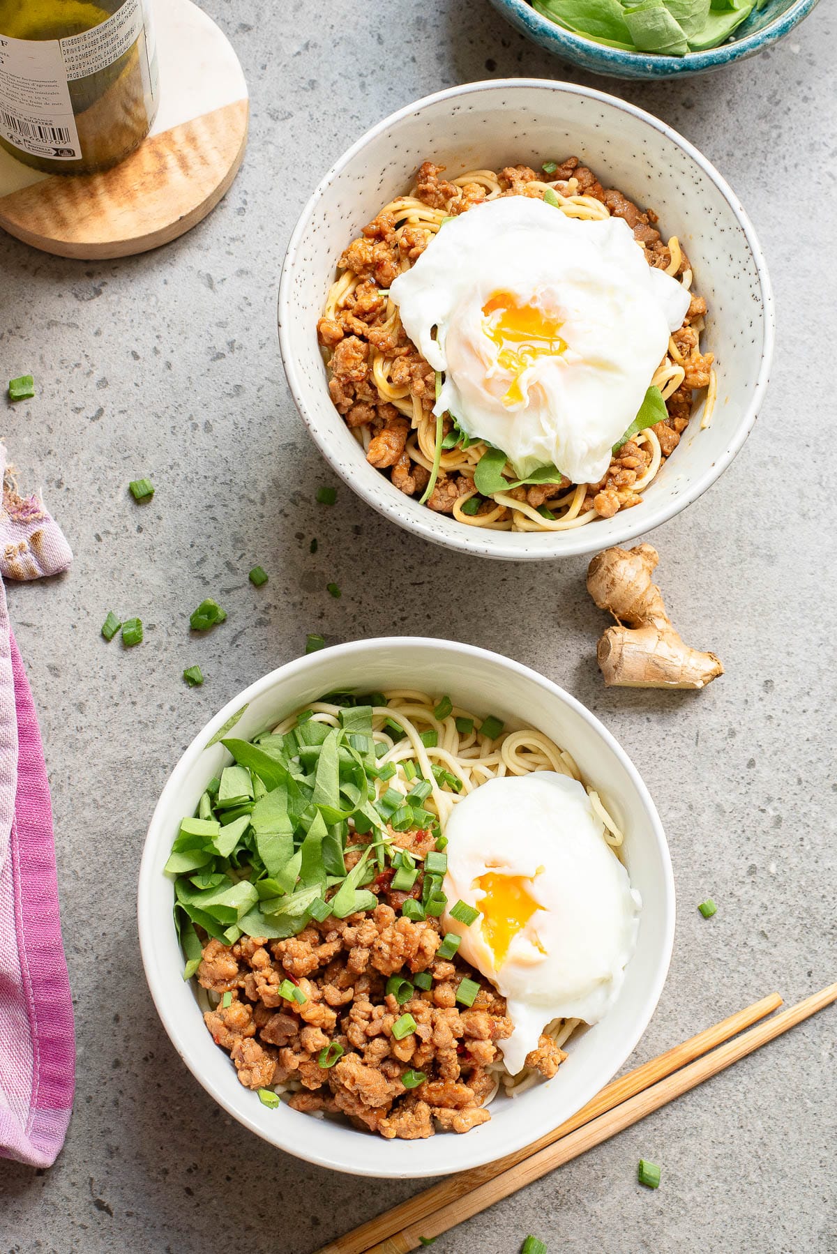 Two bowls of noodles topped with minced meat, poached eggs, and chopped greens on a gray surface. A pair of wooden chopsticks, a jar, ginger, and a towel are nearby, adding a rustic kitchen vibe to the setting.