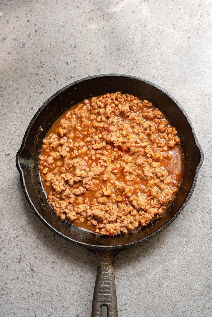A cast iron skillet filled with cooked ground meat in a rich, saucy mixture, placed on a gray surface.