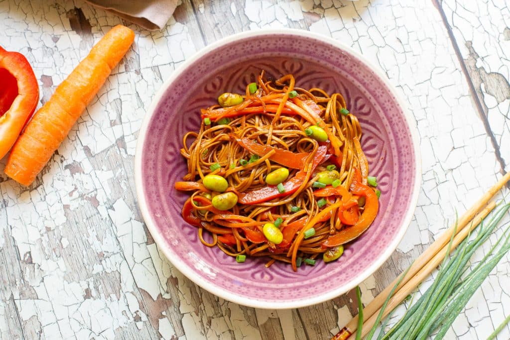 A bowl of stir-fried noodles with edamame, red bell pepper strips, and sliced green onions on a rustic wooden surface. Carrot and pepper halves are nearby, with a few chives also visible.