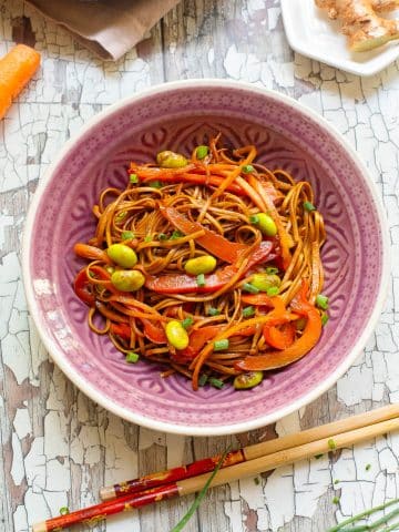 A bowl of noodles mixed with sliced red bell peppers, edamame, and green onions, garnished with sauce. Chopsticks rest on the side. Pieces of ginger and a carrot are visible in the background on a rustic white wooden surface.