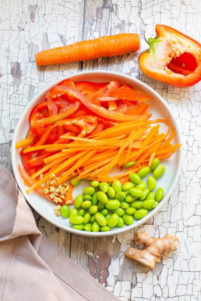 A white plate on a textured surface with sliced red bell peppers, shredded carrots, diced ginger, and edamame. Nearby are a whole carrot, ginger root, and half a red bell pepper. A beige cloth lies to the side.