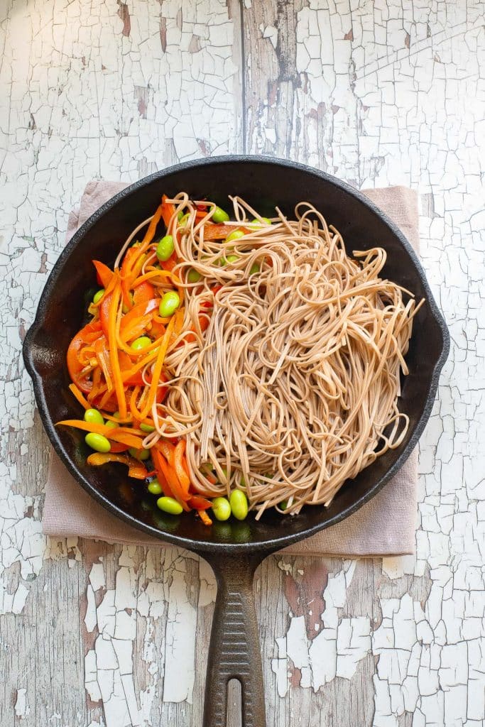 A skillet filled with soba noodles, sliced red bell peppers, and edamame on a rustic wooden surface. The noodles and vegetables are arranged neatly, showcasing a colorful, healthy dish.