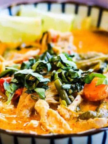 A colorful bowl of Thai red curry, featuring chicken pieces, sliced vegetables, and fresh basil leaves. The curry has a rich, creamy texture, garnished with lime wedges, and is served in a striped bowl with a spoon.