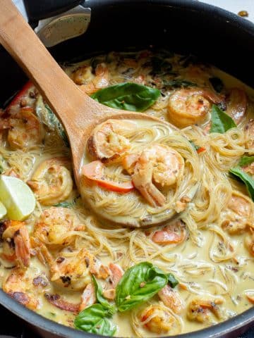 A pan filled with creamy shrimp pasta, garnished with fresh basil leaves and lime wedges. The pasta is coated in a rich, creamy sauce with visible shrimp and sliced vegetables. A wooden spoon holds a portion of the pasta above the pan.