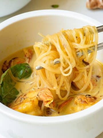 A bowl of noodle soup with chicken pieces, garnished with basil leaves. The soup has a creamy broth, and a pair of chopsticks is lifting noodles from the bowl. Ginger and garlic are in the background.