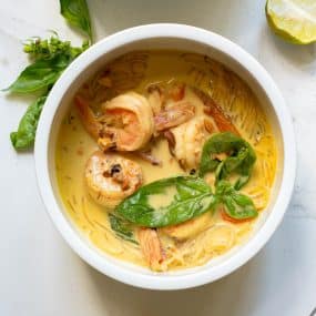 A bowl of creamy shrimp curry with noodles, garnished with fresh basil leaves. A lime wedge and more basil are on the side.