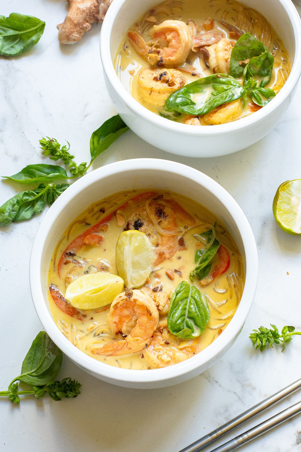 Two white bowls filled with creamy soup garnished with shrimp, lime wedges, and fresh basil leaves. The soup contains noodles and vegetables and is set on a marble surface adorned with loose basil leaves and slices of lime.