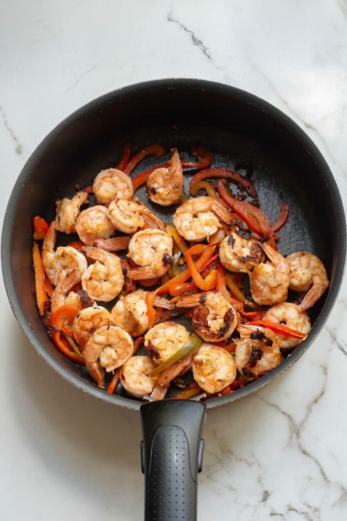 A skillet filled with cooked shrimp and sliced red bell peppers on a marble countertop.