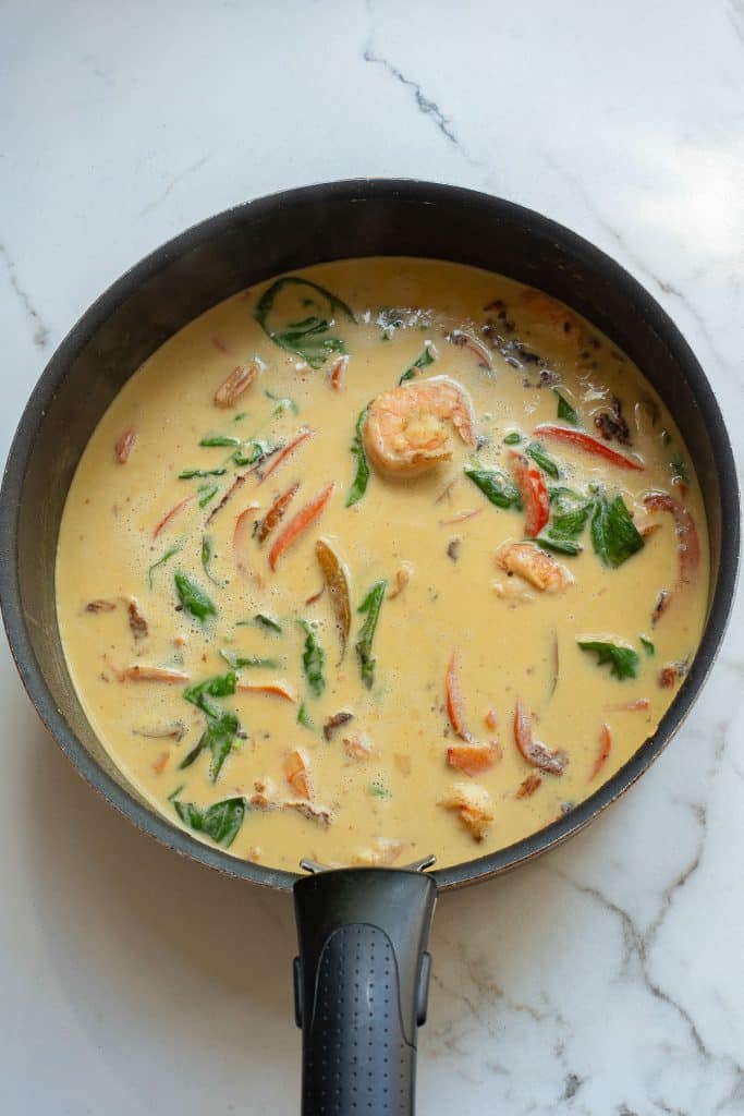 A pan filled with a creamy coconut-based curry, featuring shrimp, red peppers, spinach, and sliced mushrooms. The dish is seasoned with visible herbs and spices, creating a colorful and appetizing appearance on a marble countertop.