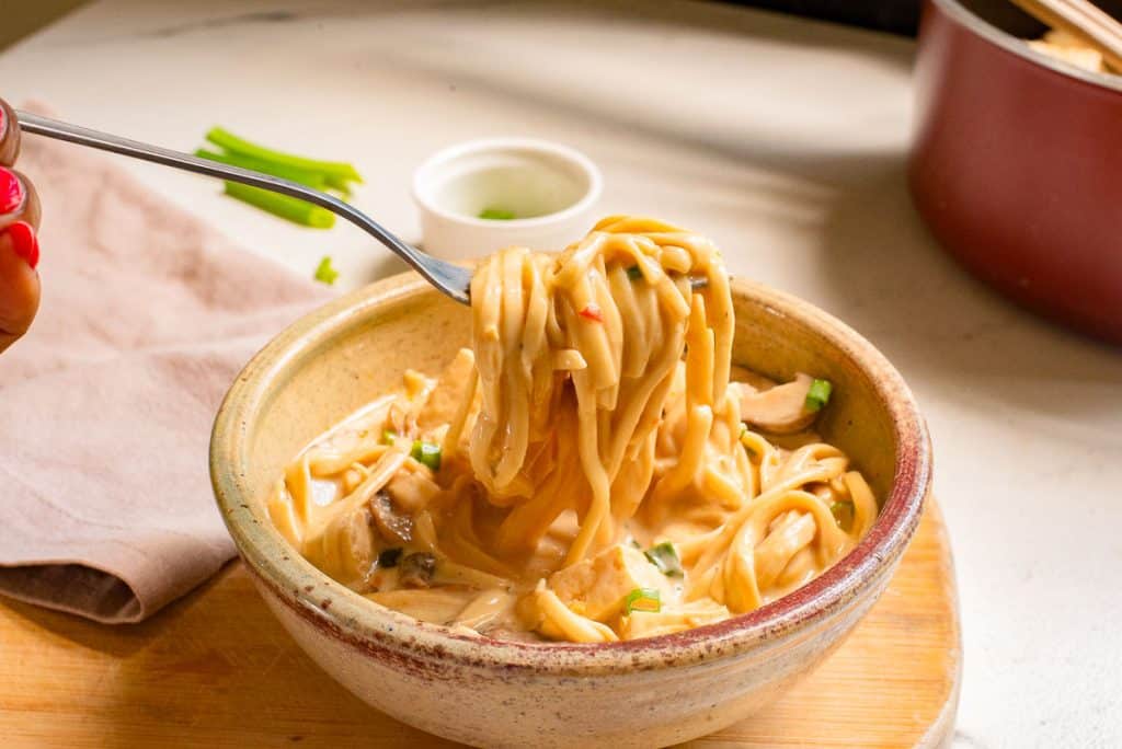 A person holding a fork twirls creamy noodles from a bowl. The dish is garnished with sliced green onions. The setting includes a napkin, a small cup, and a pot in the background, all on a wooden surface.