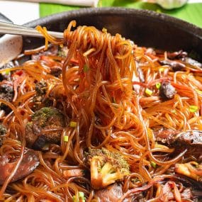 A close-up of chopsticks picking up saucy glass noodles from a pan. The noodles are mixed with broccoli, mushrooms, and sliced vegetables, all coated in a rich, dark sauce. The dish is vibrant and appetizing.