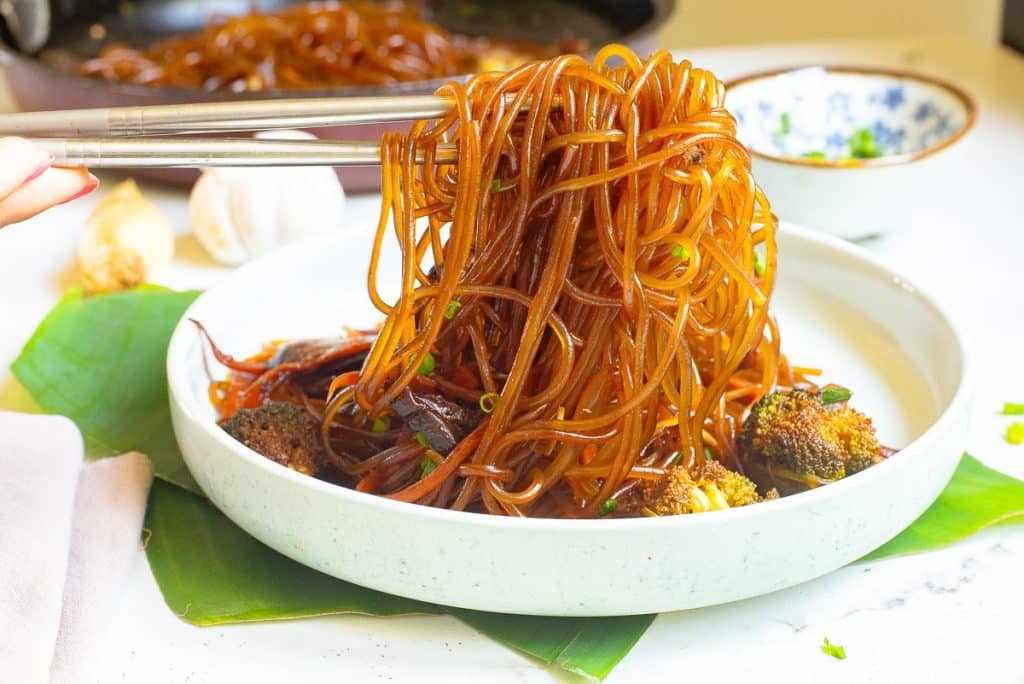 A plate of soy sauce noodles is held with chopsticks. The noodles are garnished with slices of mushroom and broccoli, set on a white surface with a small bowl in the background.