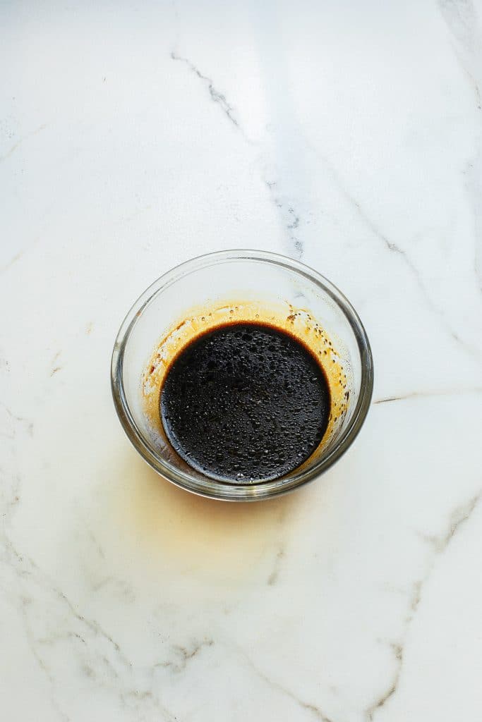 A small glass bowl with dark, thick liquid sits on a white marbled surface. The liquid has a frothy texture on top, resembling a concentrated sauce or coffee extract.