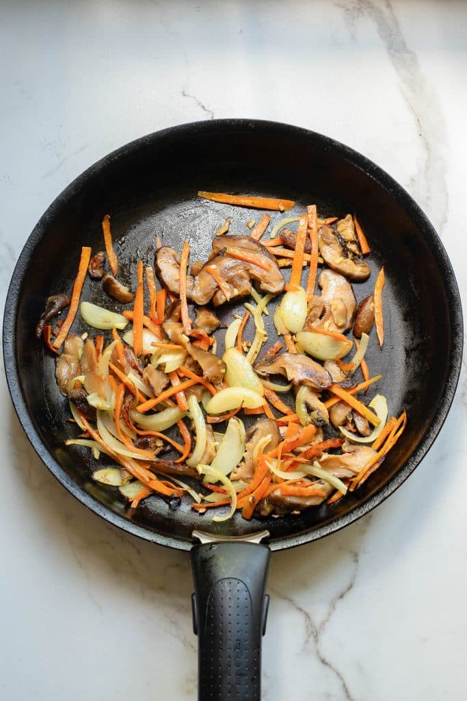 A black pan holds sautéed vegetables, including sliced mushrooms, carrots, and onions, on a light marble surface. The vegetables are lightly browned, suggesting they are being cooked.