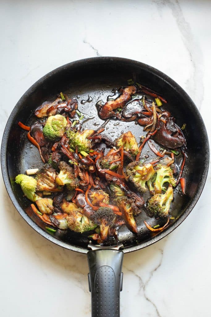 A black frying pan on a marble surface contains sautéed vegetables, including broccoli, carrots, mushrooms, and onions. The vegetables are lightly browned, seasoned, and glistening.
