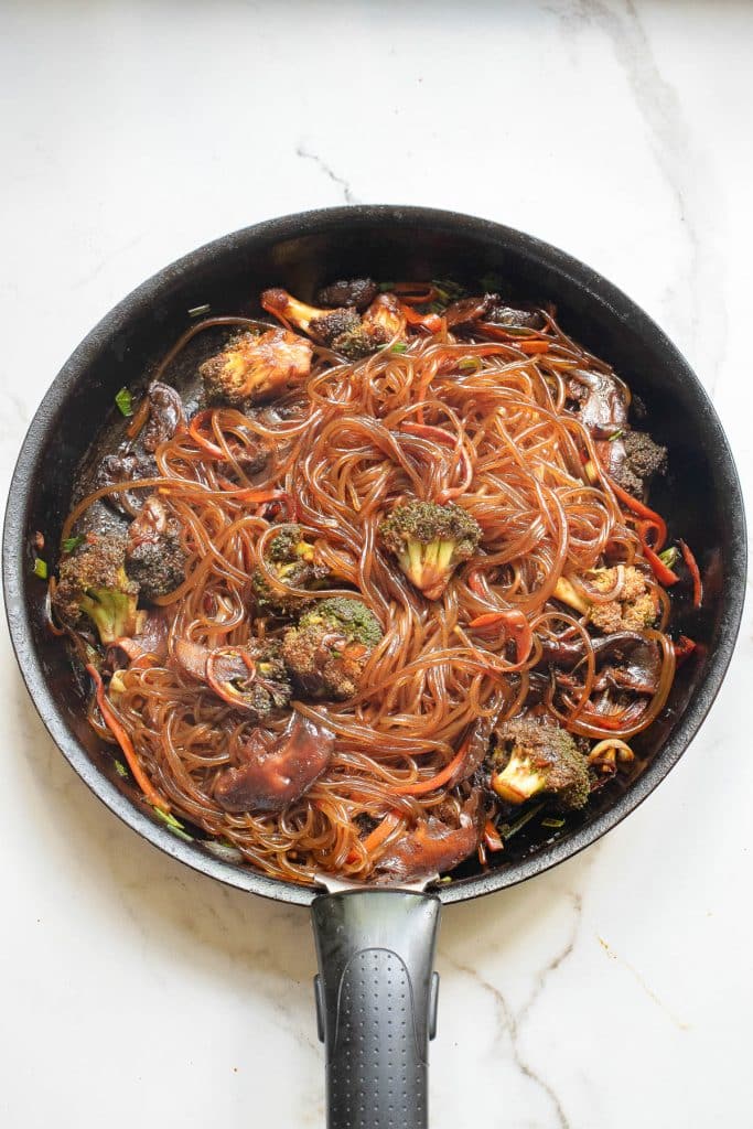 A skillet filled with cooked noodles, broccoli, and carrots in a dark, glossy sauce, placed on a light marble countertop.