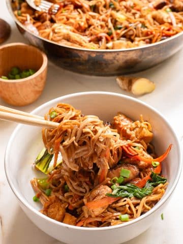 A close-up image of a bowl filled with stir-fried noodles, chicken pieces, bok choy, and carrots. A fork is lifting a portion of the noodles from the bowl. A skillet with more noodles and a small bowl of chopped green onions are visible in the background.