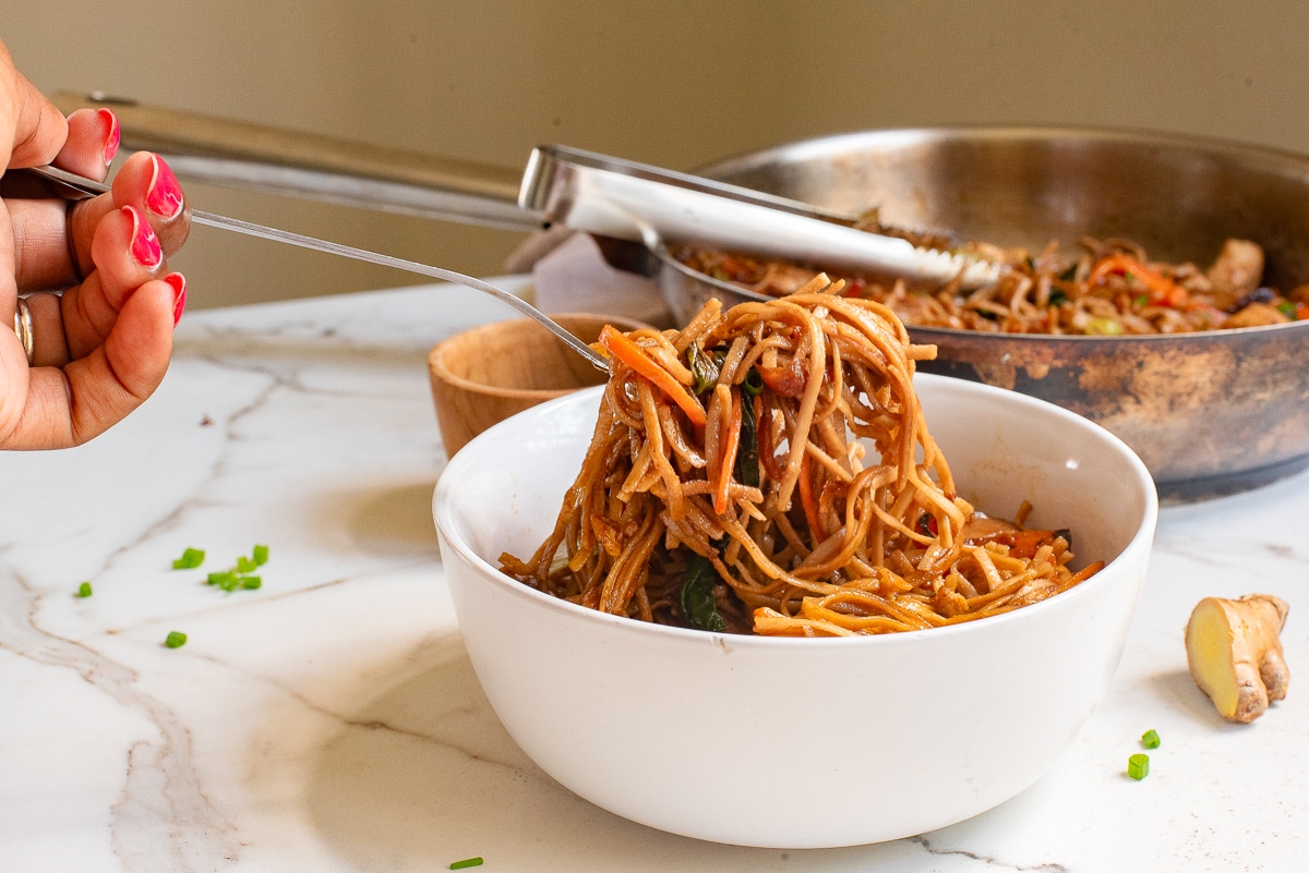 A person is serving a portion of vegetable stir-fried noodles from a pan into a white bowl with a fork. The marble countertop has chopped chives and a ginger piece. A wooden bowl and tongs are nearby.