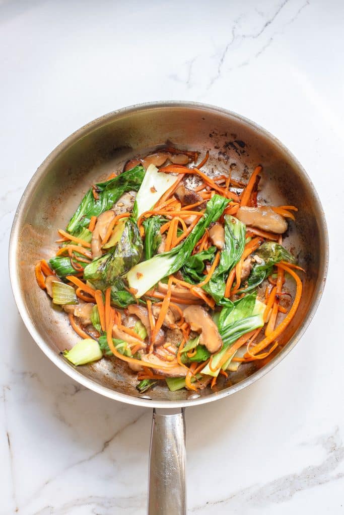 A stainless steel pan on a marble surface contains a stir-fry with green bok choy, orange carrot strips, and brown mushrooms. The vegetables are lightly covered in a sauce.
