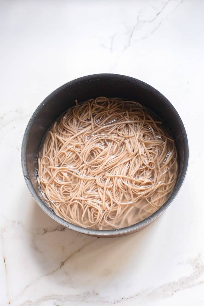A black pot filled with cooked spaghetti noodles, soaking in cloudy water, sits on a white marbled countertop. The noodles are tangled together, and steam rises from the pot, indicating they are hot.