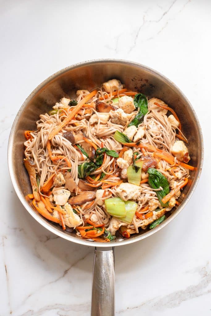 A stainless steel frying pan contains a colorful stir-fry. The dish includes soba noodles, leafy greens, carrots, mushrooms, and chunks of tofu, all mixed with a light sauce. The pan is placed on a white marble countertop.