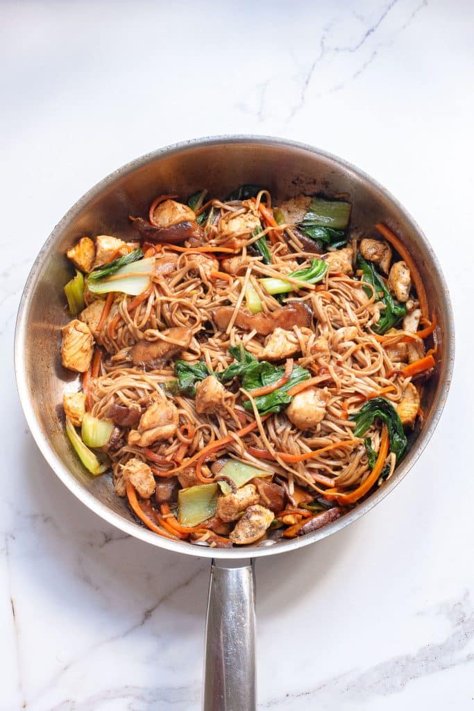 A silver pan filled with cooked stir-fry noodles mixed with pieces of chicken, bok choy, carrots, and other vegetables. The dish appears well-seasoned and colorful, resting on a white marble surface.