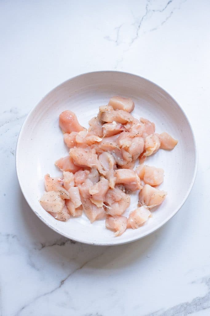 A white bowl on a marble countertop contains small, raw chicken pieces seasoned with salt and pepper. The pink and pale yellow chicken pieces are irregularly cut and appear ready for cooking.