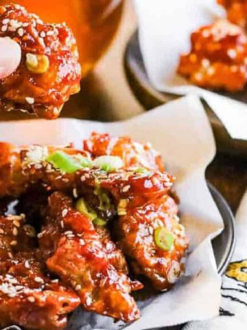 A person holding up a piece of glazed fried chicken garnished with sesame seeds and green onions. More pieces are served on a plate lined with parchment paper, with a decorative napkin nearby.