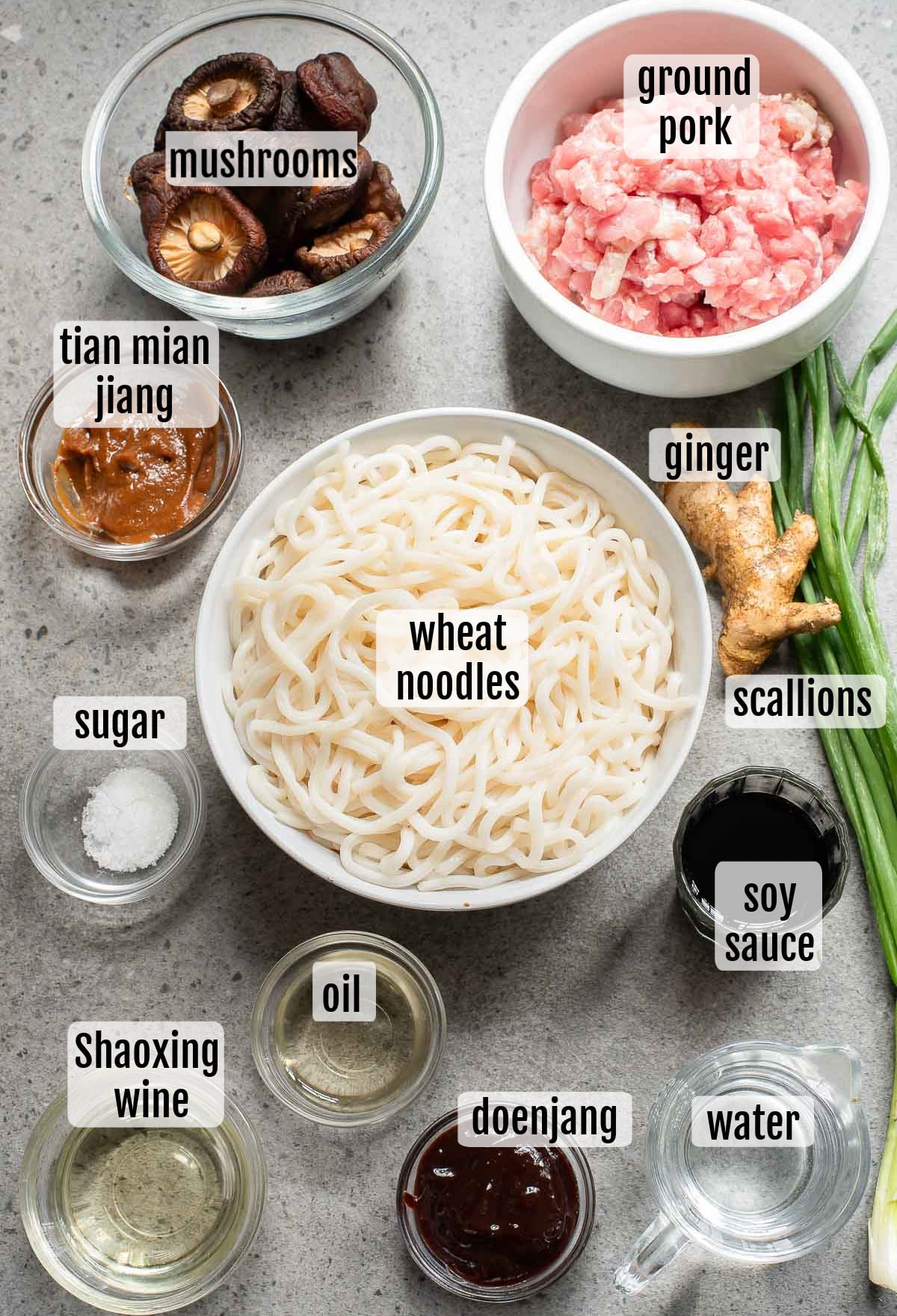 The ingredients for Beijing noodles are meticulously arranged on a table: bowls of mushrooms, ground pork, wheat noodles, ginger, and scallions. Nearby sit containers of sugar, tian mian jiang, oil, soy sauce, Shaoxing wine, doenjang, and water. Each item is clearly labeled for easy preparation.