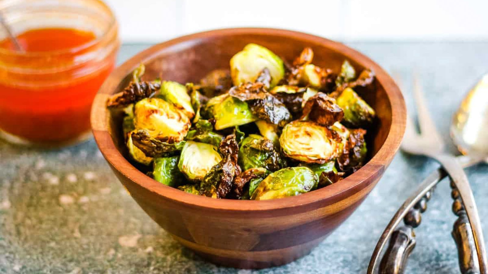 A wooden bowl filled with roasted Brussels sprouts. A jar of red sauce and a pair of tongs are in the background.