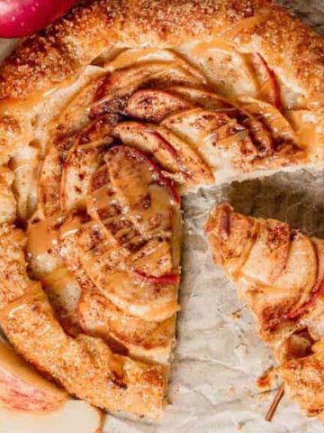 A freshly baked apple galette with a golden crust sits on parchment paper atop a cooling rack. One slice is cut and partially removed. Surrounding the galette are whole red apples and cinnamon sticks.