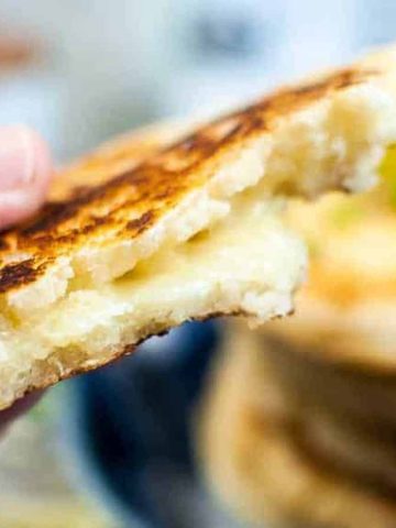 A hand holding a cheesy arepa with a bite taken out, revealing the gooey cheese filling. In the background, there is a stack of arepas on a plate, slightly blurred.