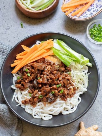 A black bowl filled with noodles topped with minced meat, diced vegetables, and sprouts. Sliced cucumber and carrot sticks are arranged on the side. Additional carrot sticks and cucumber slices are in small bowls nearby. A piece of ginger is on the table.